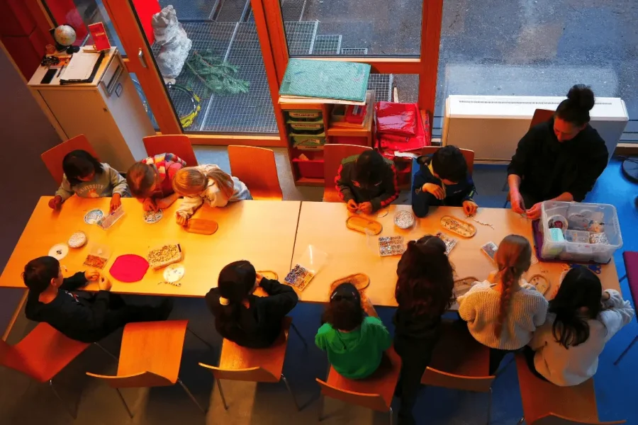 Eine Gruppe Kinder sitzt um einen Tisch und arbeitet an Kunst- und Bastelprojekten, wobei ein Erwachsener ihr hilft, wie von oben zu sehen ist.