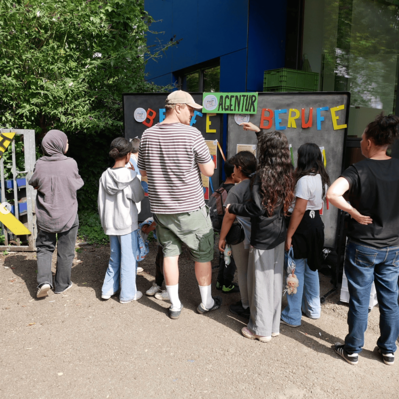 Eine Gruppe von Kindern und zwei Erwachsene stehen im Freien vor einer Informationstafel mit der bunten Aufschrift „AGENTUR BERUFE“ und schauen sich gemeinsam die dort angebrachten Aushänge an.