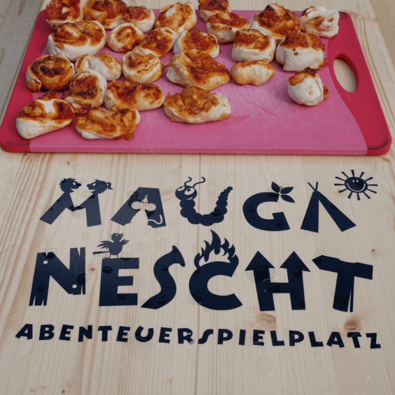 Pizzaschnecken auf einem roten Schneidebrett. Darunter steht der Schriftzug "Mauga Nescht Abenteuerspielplatz"