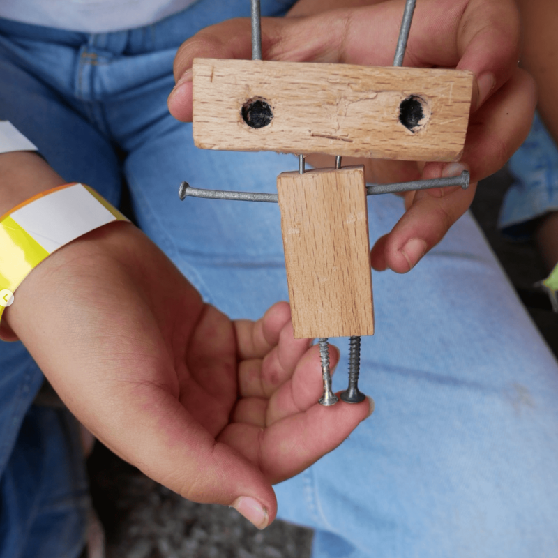 Nahaufnahme von Kinderhänden, die einen kleinen, handgemachten Holzroboter halten. Die Figur besteht aus 2 Holzklötzchen mit gebohrten Augenlöchern sowie Nägeln und Schrauben, die als Arme, Beine und Antennen dienen.