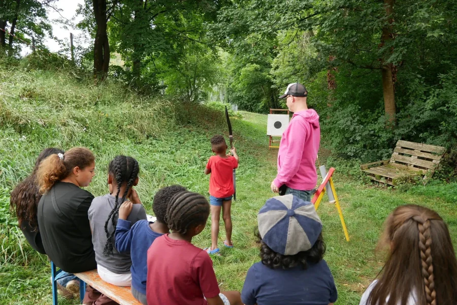Kinder beobachten, wie ein Kind unter Anleitung eines Erwachsenen mit Pfeil und Bogen auf eine Zielscheibe schießt.