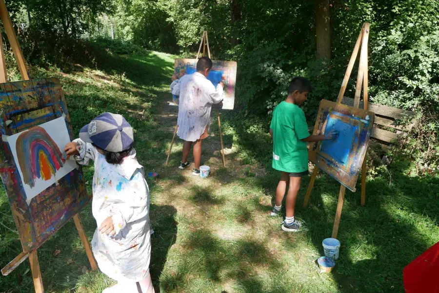 Kinder malen draußen im Grünen an Staffeleien mit Farben und Pinseln.