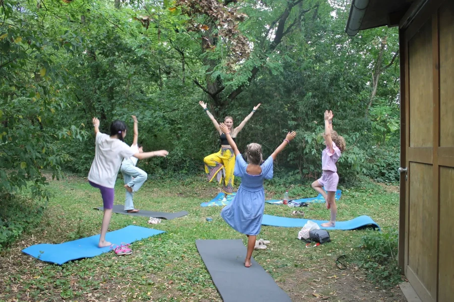 Eine Yogagrupe, bestehend aus einer Yogalehrerin und vier Mädchen, übt die Baumpose im Freien auf Matten, umgeben von dichtem Grün.